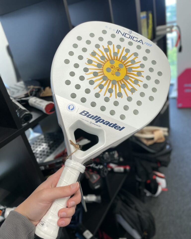 Käsi pitelee valkoista Bullpadel-mailaa, takana myymälän hyllyt — kokeile ennen ostoa